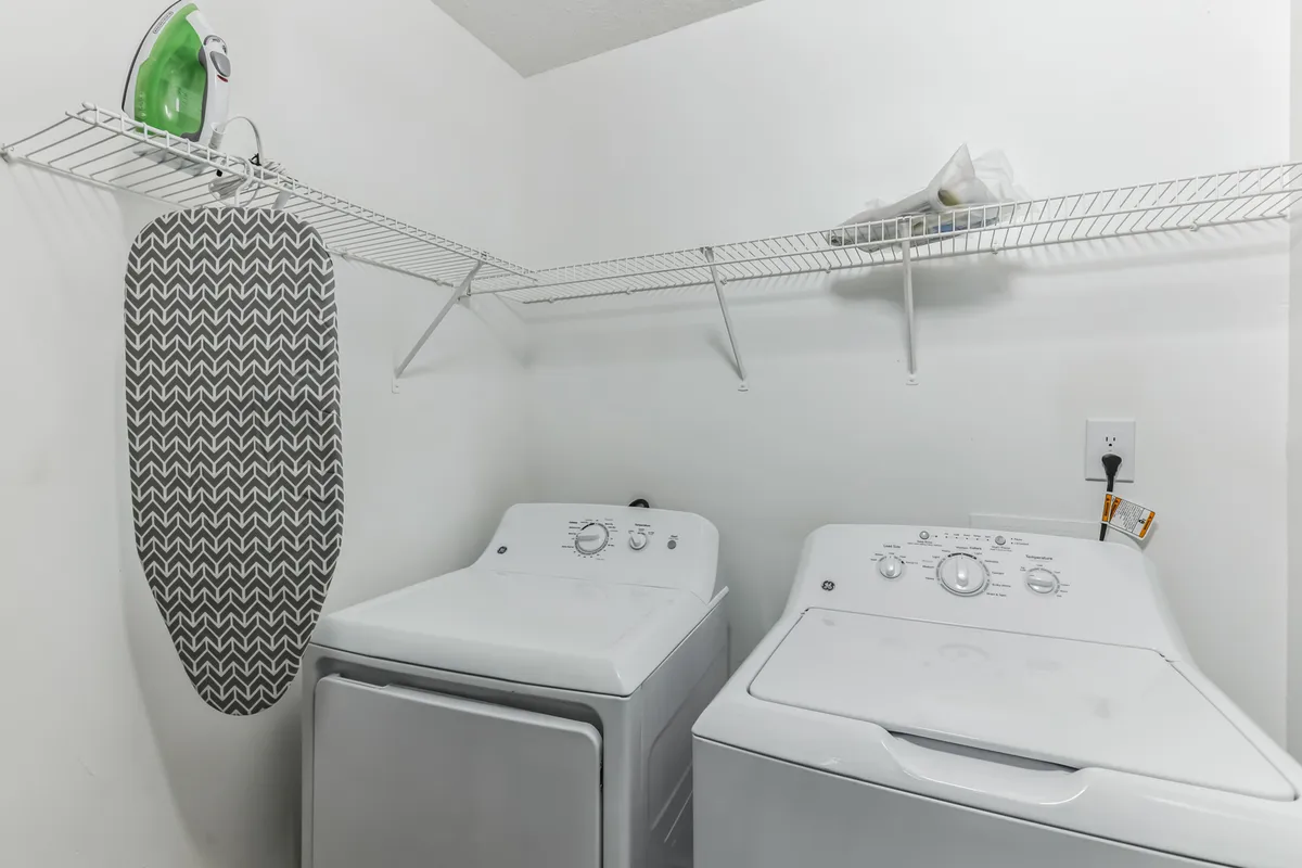 A focused view of the laundry area, featuring a modern washing machine, dryer, ironing board, and iron. The setup illustrates the practical and accessible arrangement for handling laundry tasks within the apartment.