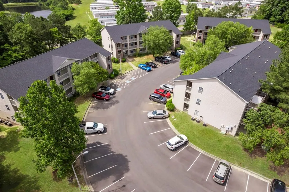 A broad view of the apartment buildings with a parking area in the foreground and abundant green trees in the background. The scene is bright and friendly, showcasing the external environment where residents gather, children play, and vehicles are easily parked.