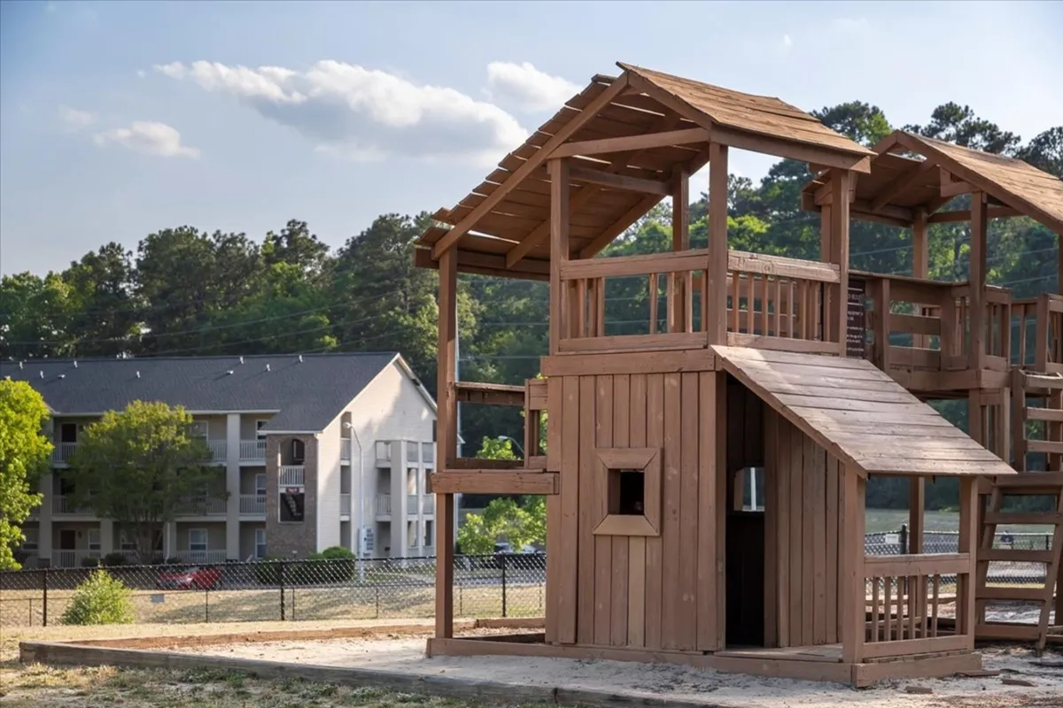 This exterior image features a lively playhouse structure surrounded by mature trees, with an apartment building visible in the background. Children are seen playing and families are gathered, creating a scene of community and outdoor enjoyment.