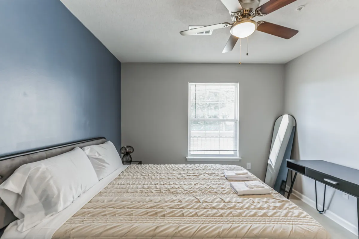 A serene bedroom scene with a neatly made bed, a window letting in natural light, a sleek mirror, and a minimalist desk. The space exudes comfort, organization, and the versatile functionality of the apartment’s interior.
