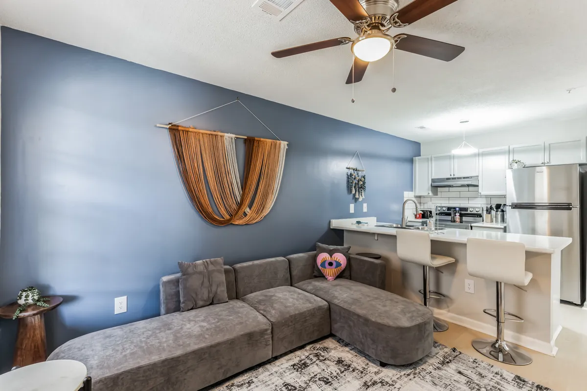 The hero image showcases a modern, open-concept living space featuring a plush sofa adorned with decorative pillows, a stylish kitchen island, and chic wall art. The room feels comfortable and inviting, perfect for relaxing after a day exploring Fayetteville. Subtle design touches and ample natural light create a welcoming atmosphere.