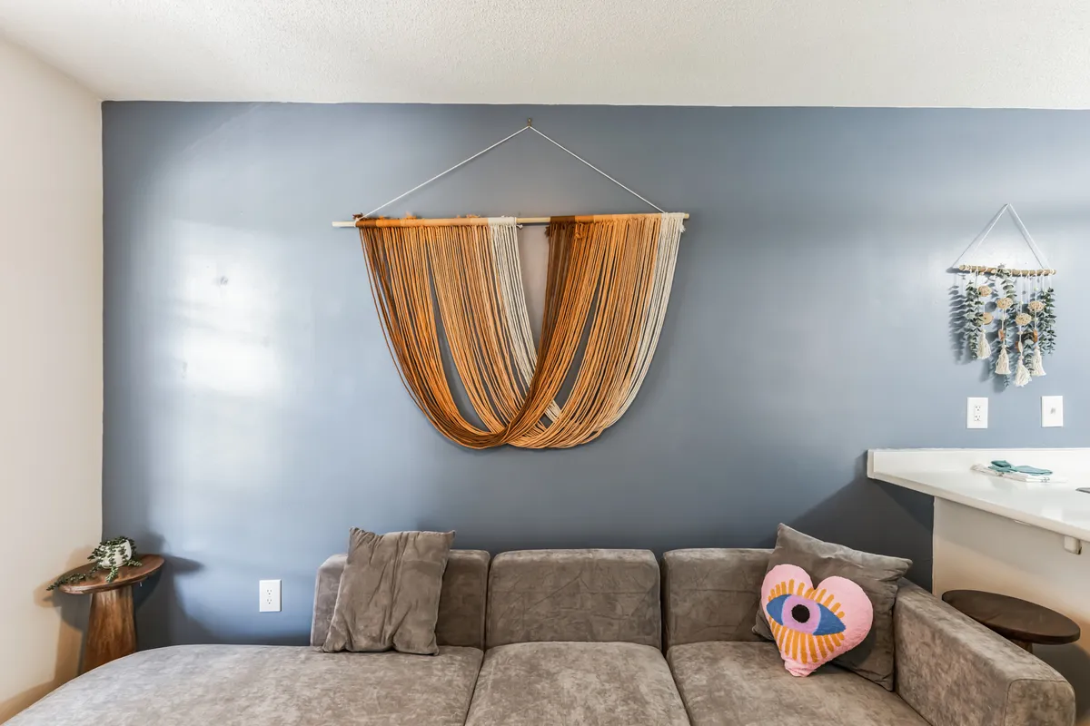 A soft couch with a colorful pillow sits against a textured wall hanging, accompanied by a wooden side table. The space exudes creativity and comfort, perfect for unwinding or intimate conversations, and is filled with natural light.