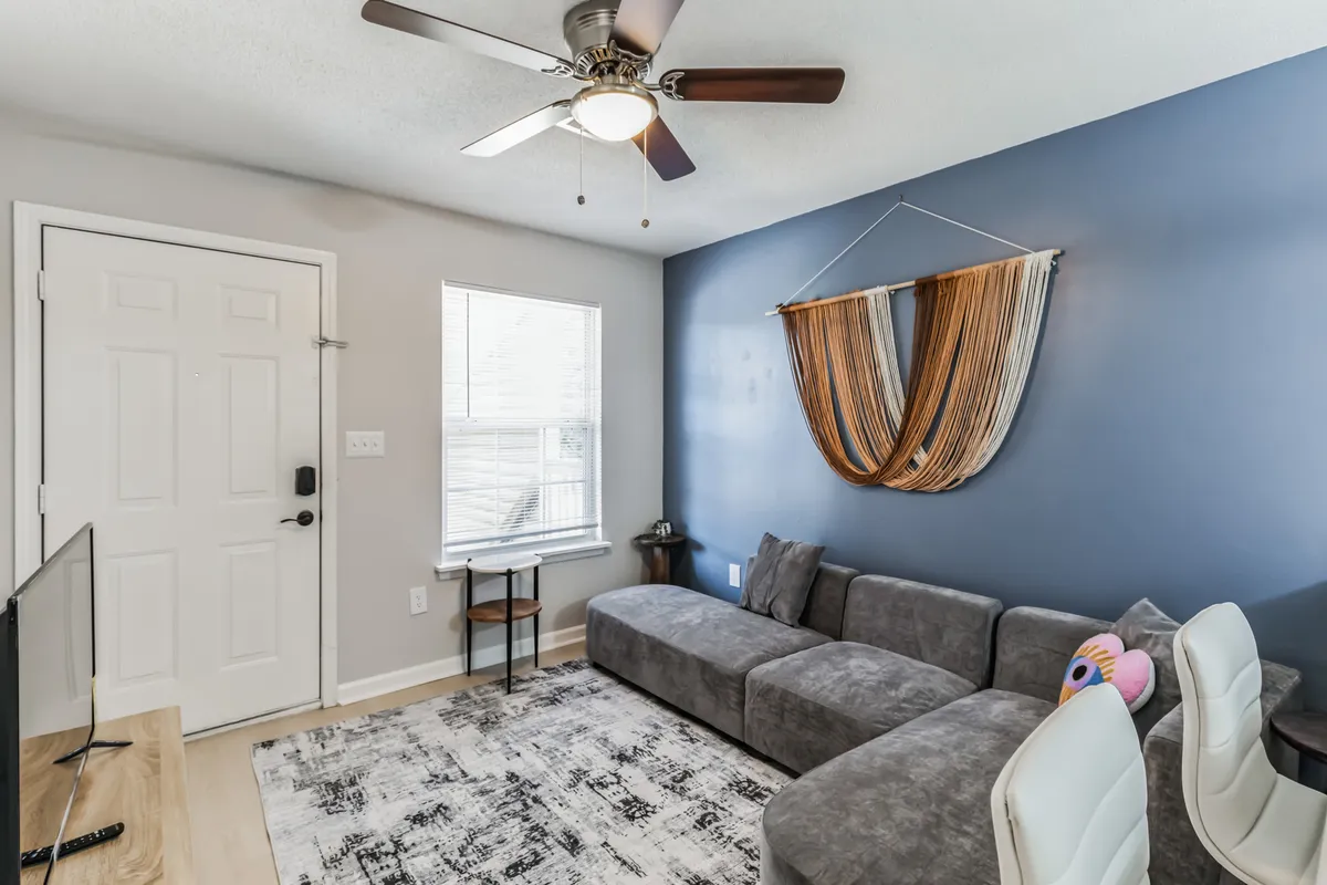 A welcoming living room featuring a plush sofa, tasteful wall decor, a sleek coffee table, a large window that brings in ample natural light, and a TV for entertainment. The room is styled for both relaxation and socializing, giving a sense of homeliness and warmth.