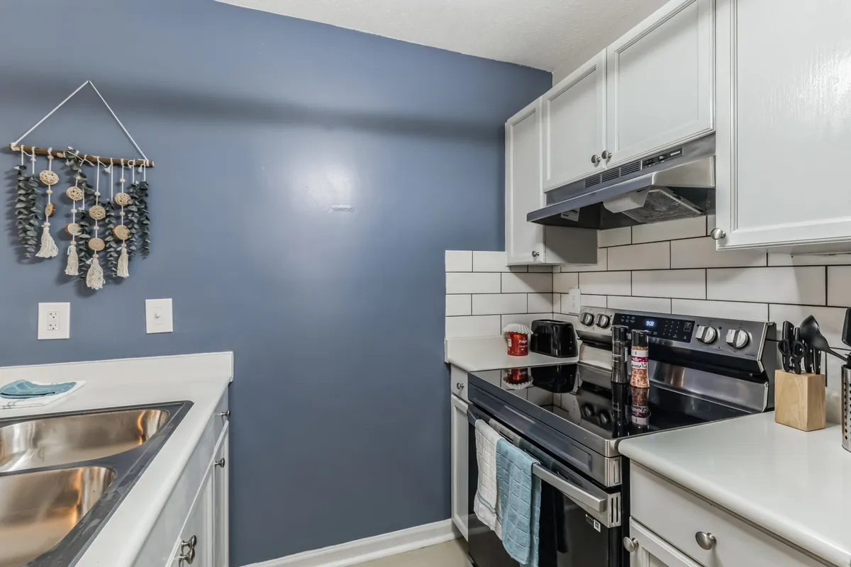 A bright, welcoming kitchen featuring stainless steel appliances, elegant white cabinetry, a blue accent wall, and decorative hanging pieces. The space is arranged for easy cooking and casual dining, evoking a sense of warmth and homeliness.