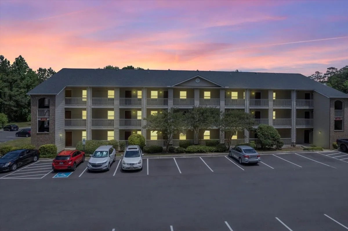 This image features a multi-story apartment building with glowing windows set against a colorful sunset sky. Residents can be seen relaxing on balconies and socializing in the parking lot below, enjoying the tranquil evening atmosphere.