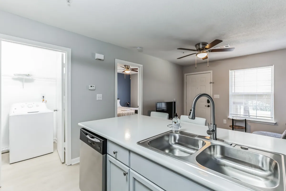 This image showcases a bright and welcoming open-concept living space that features a kitchen island, living area, large windows allowing natural light, and—critically for this section—a washer and dryer unit in the background. The scene captures a blend of functional amenities within a comfortable, home-like setting.