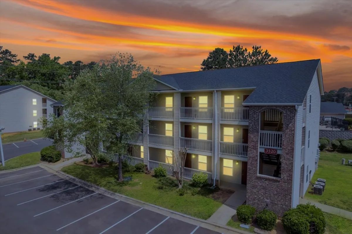 Captured at dusk, this scene shows an apartment building framed by surrounding trees and a soft sunset sky. Residents are visible on balconies and in the parking lot, suggesting a calm, communal vibe ideal for evening relaxation.
