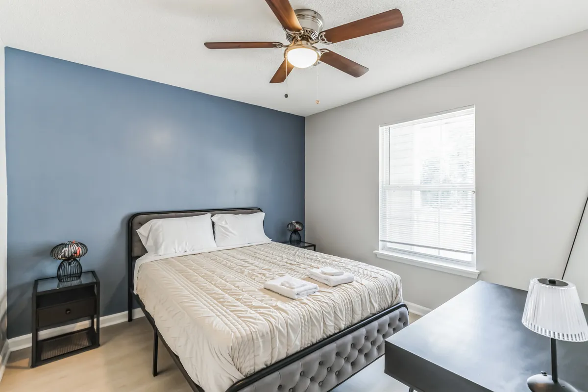This image showcases a serene bedroom featuring a plush queen bed dressed in soft linens, a nightstand with gentle lighting, a large window allowing natural light to fill the space, and a ceiling fan overhead. The atmosphere is calm and inviting, ideal for relaxation and restful sleep.