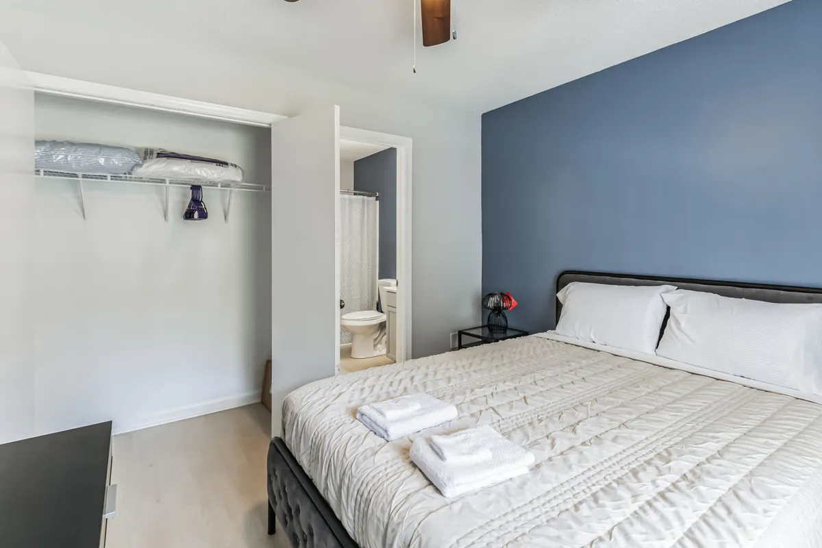 A tranquil bedroom scene showing a neatly made bed, a closet providing convenient storage, a bathroom door for private access, and a decorative lamp that adds a touch of coziness. The space feels organized and restful, supporting both relaxation and practical needs.