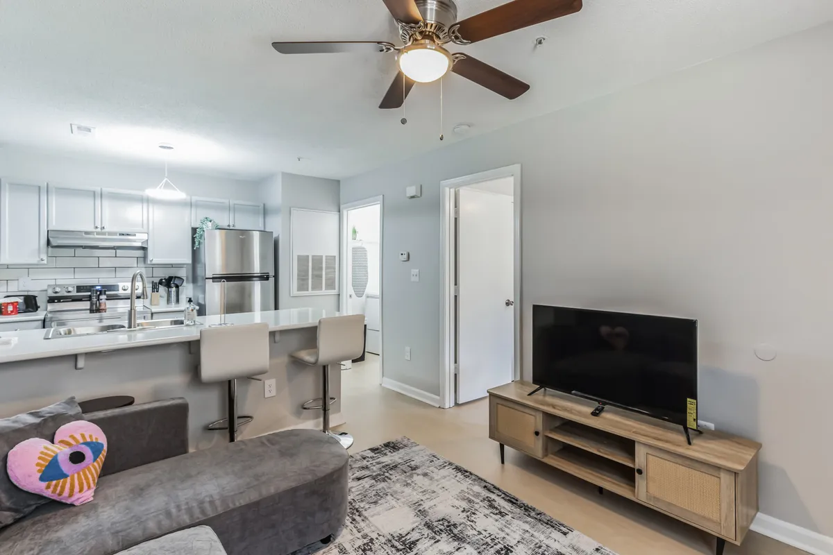 A cozy interior view with a soft sofa accented with a decorative pillow, a modern kitchen area in the background, and a TV unit. The space feels inviting and designed for comfort, relaxation, and casual entertainment.