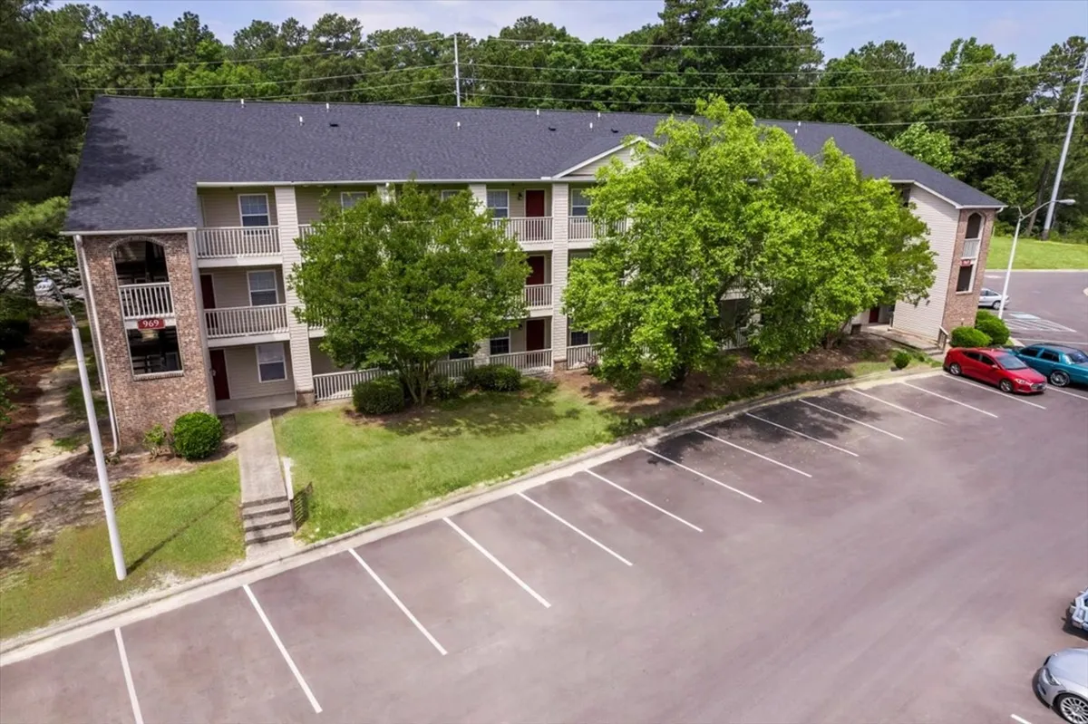 This exterior shot captures the well-maintained facade of Carlson Bay, surrounded by lush green trees and featuring spacious balconies overlooking a peaceful parking lot. The image conveys a sense of welcoming residential living and hints at the ease of coming and going for busy military personnel and families.